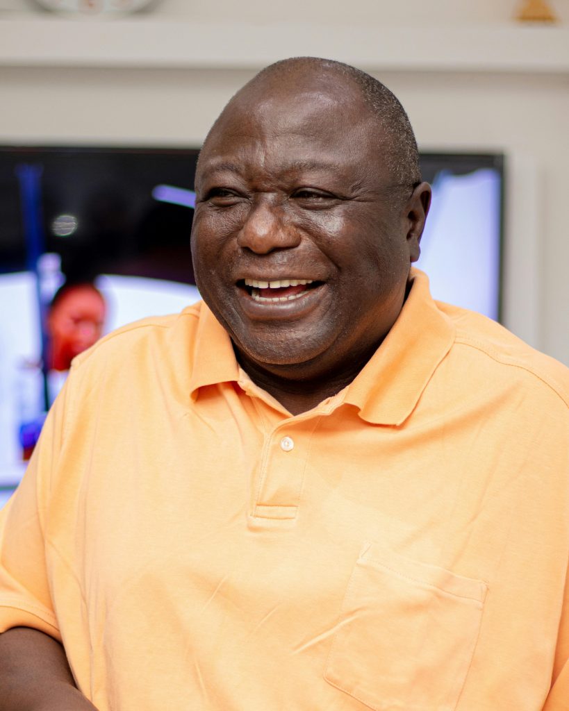 African man wearing orange polo shirt smiling warmly in an indoor setting. Captured in Nigeria.