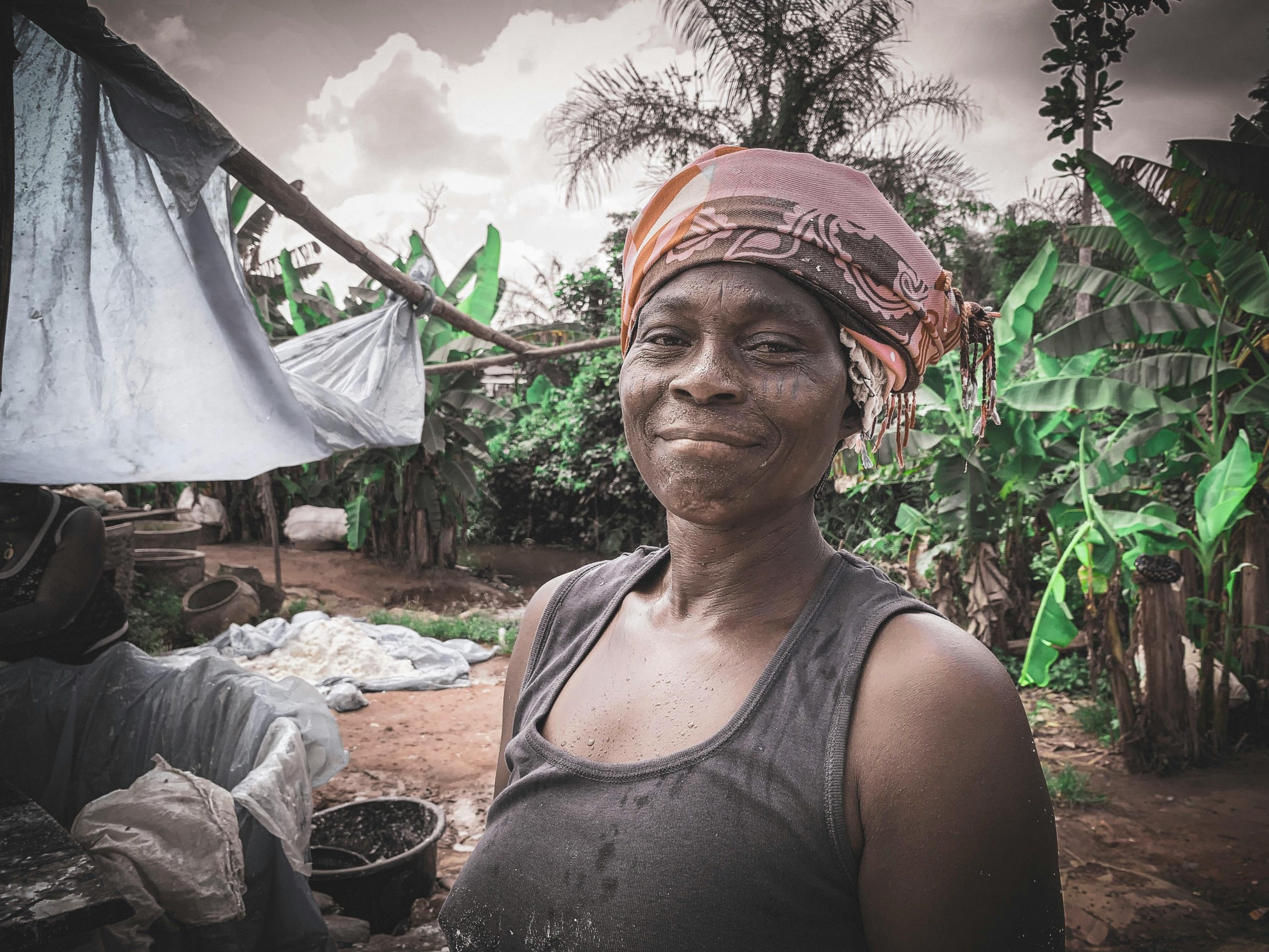 A smiling woman in rural Nigeria, showcasing traditional lifestyle with lush green background.