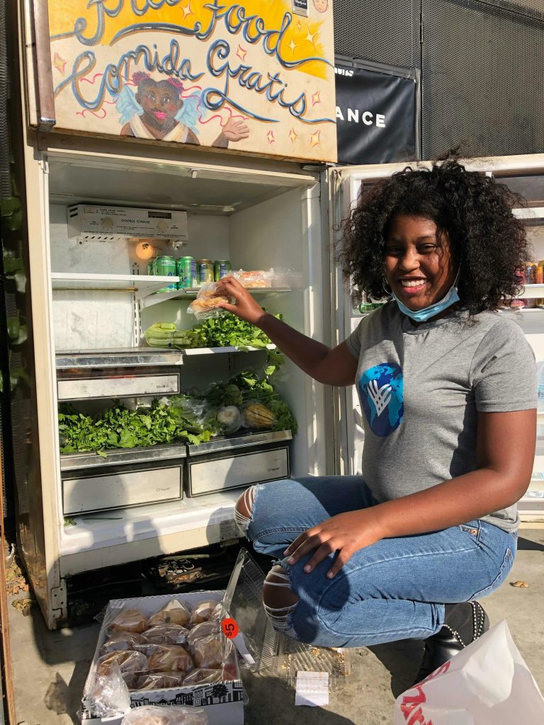 Smiling woman stocks a community fridge with fresh greens and groceries outdoors.